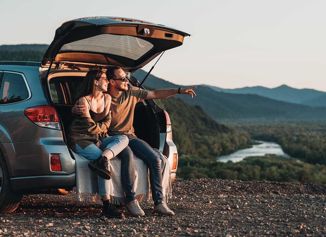 Contact - Young Couple Sitting in the Open Back Trunk of an SUV While Looking Out at the Mountain Landscape Around Them at Sunset